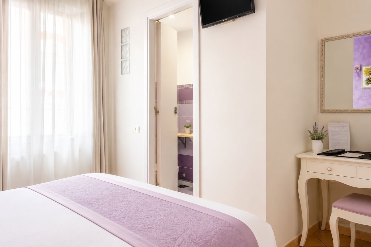 Habitación de hotel con cama, escritorio, silla, televisor y baño visible a través de una puerta abierta.