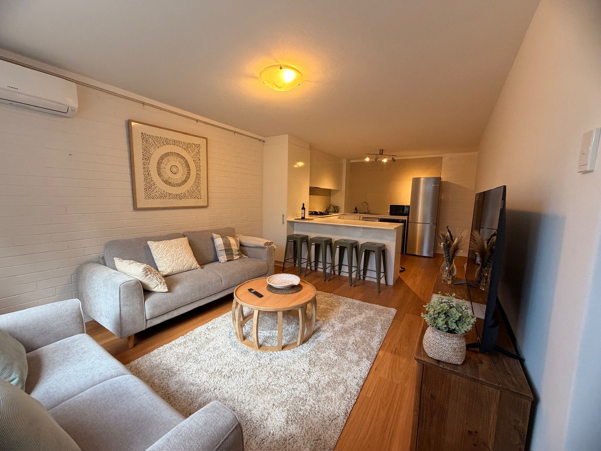 A modern living room with a sofa, coffee table, and a TV. There is a kitchen area with a fridge and a dining table.