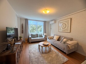 A living room with a sofa, a coffee table, a TV, and a window with curtains. - Newly Renovated Coastal Stay in Mosman Park, 2 Br (Mosman Park)