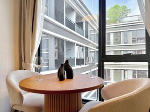 A room with a round wooden table, two chairs, a window with a view of a building, and a pair of vases. - Serene 1-Bedroom Apartment with Pool & Gym in Khlong Toei (Khlong Toei)