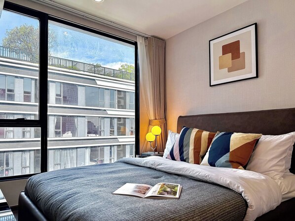 A hotel room with a large bed, a nightstand, a lamp, a framed picture on the wall, and a view of a building outside. - Serene 1-Bedroom Apartment with Pool & Gym in Khlong Toei (Khlong Toei)