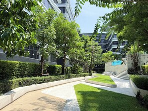 Uma área externa ajardinada, com caminhos, vegetação e edifícios modernos ao fundo.