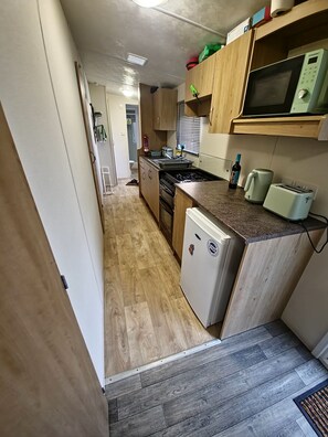 A compact kitchen with wooden cabinets, a built-in microwave, a gas stove, and a small refrigerator. - CocoTony Cosy 3 bed caravan (Abergele)