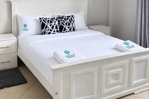 A white bed with a headboard, two pillows, a white nightstand, a drawer unit, and a towel with a logo. - Likizo Homes - Rehema Apartment, 202, Naivasha (Naivasha)