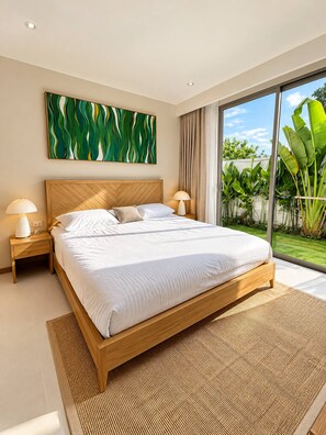 A modern bedroom with a large bed, wooden headboard, and a view of lush greenery through a sliding glass door. - Brand New Private 3-Bedroom Pool Villa in Bang Tao, Phuket (Thep Krasatti)