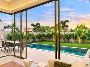A modern outdoor pool area with a glass wall, a dining table, and chairs. - Brand New Private 3-Bedroom Pool Villa in Bang Tao, Phuket (Thep Krasatti)