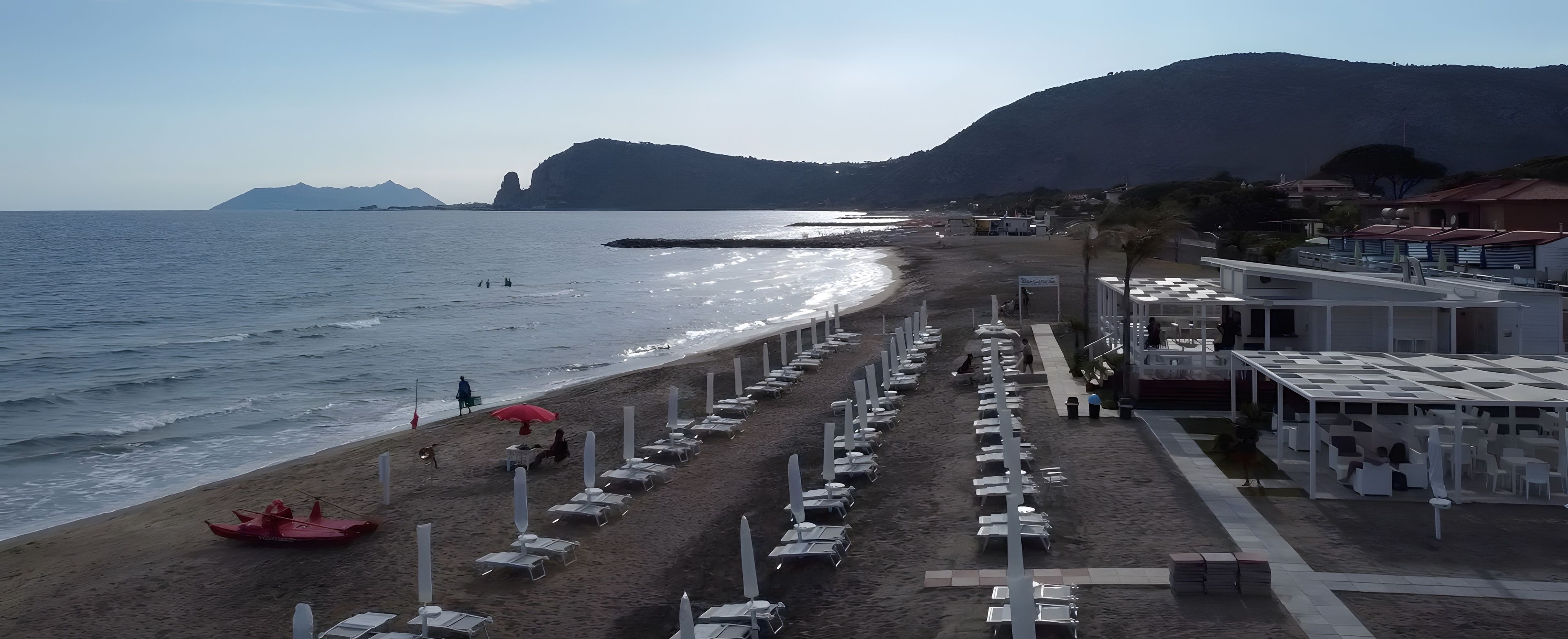 Ein Hotel direkt am Strand mit Reihen von Liegestühlen und Sonnenschirmen, ein Strand und ein Berg im Hintergrund.