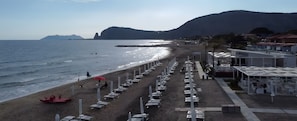 Een hotel aan het strand met rijen ligbedden en parasols, een zandstrand en een berg op de achtergrond.