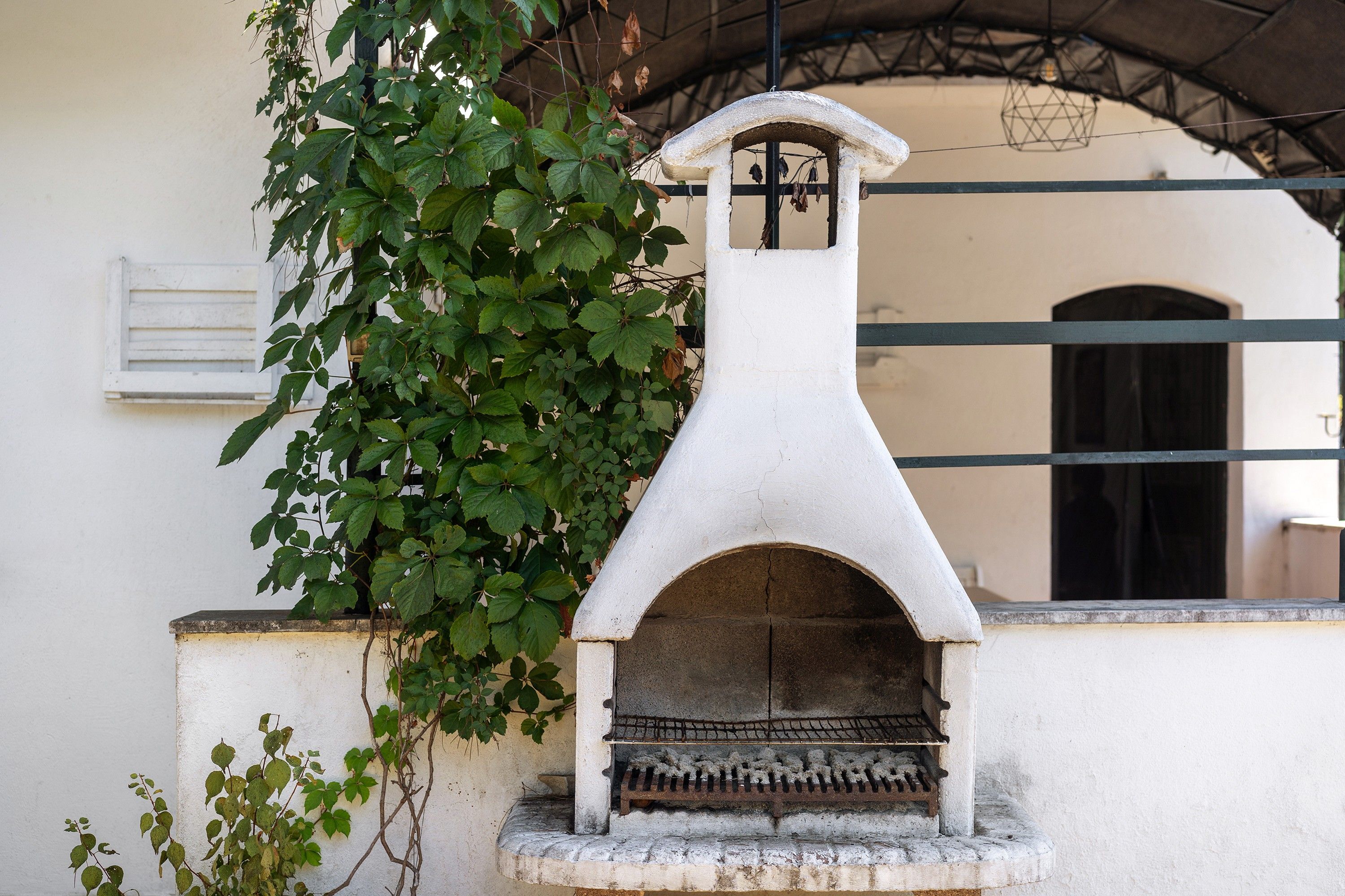 Un horno de piedra al aire libre con un diseño sencillo y funcional, frente a una pared blanca con una ventana y una reja de metal al fondo.