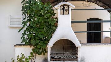 Un horno de piedra al aire libre con un diseño sencillo y funcional, frente a una pared blanca con una ventana y una reja de metal al fondo.