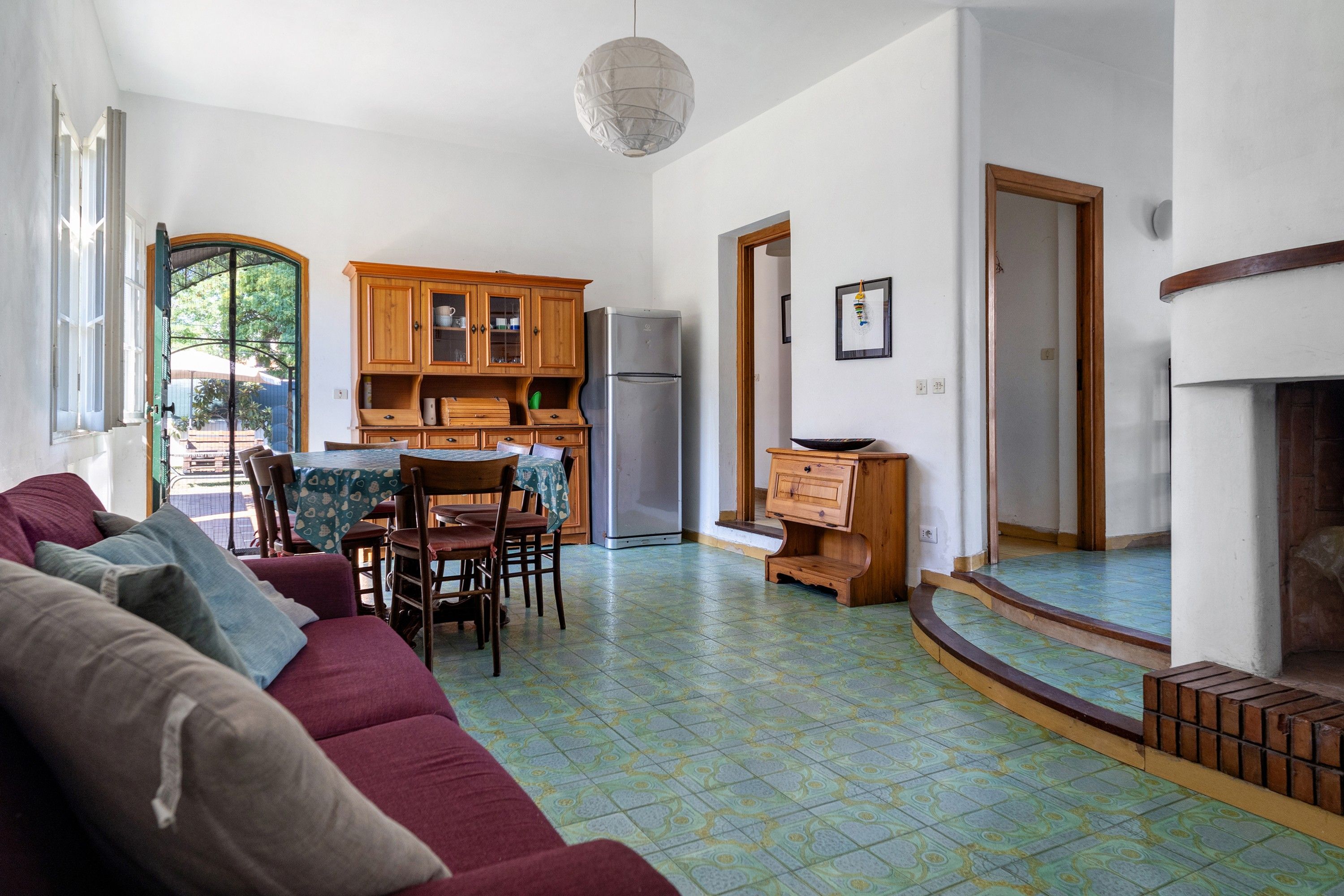 A living room with a sofa, chairs, a dining table, wooden cabinets, a refrigerator, and a patterned floor.