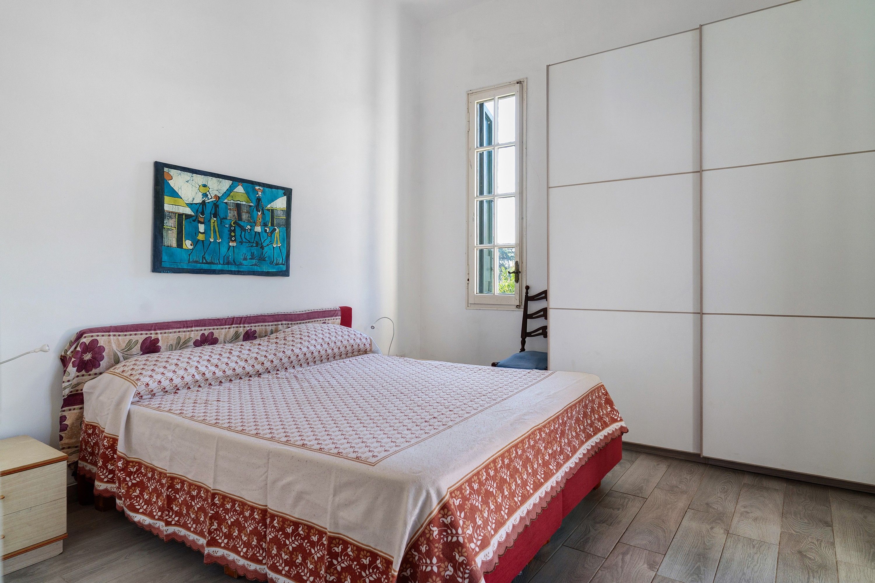 A bedroom with a bed, a wardrobe, a window, and a painting on the wall.