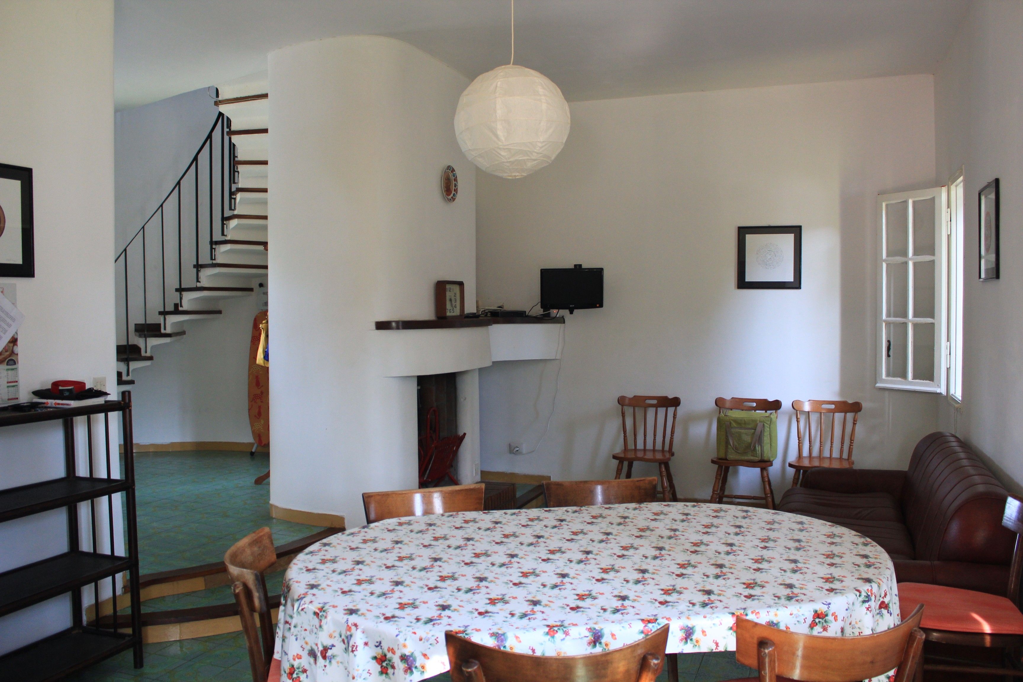 Ein Essbereich mit einem runden Tisch, der mit einer blumigen Tischdecke bedeckt ist und von Holzsesseln umgeben wird. Im Hintergrund ist eine Treppe zu einer oberen Ebene zu sehen.