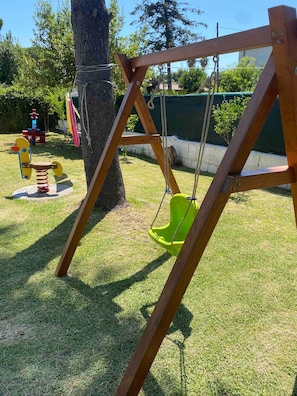 Un columpio de madera con asiento verde, un árbol y juegos infantiles al fondo.
