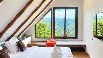A bedroom with a sloped ceiling, a large window with a view of greenery, a bed with pillows, and a bedside table.