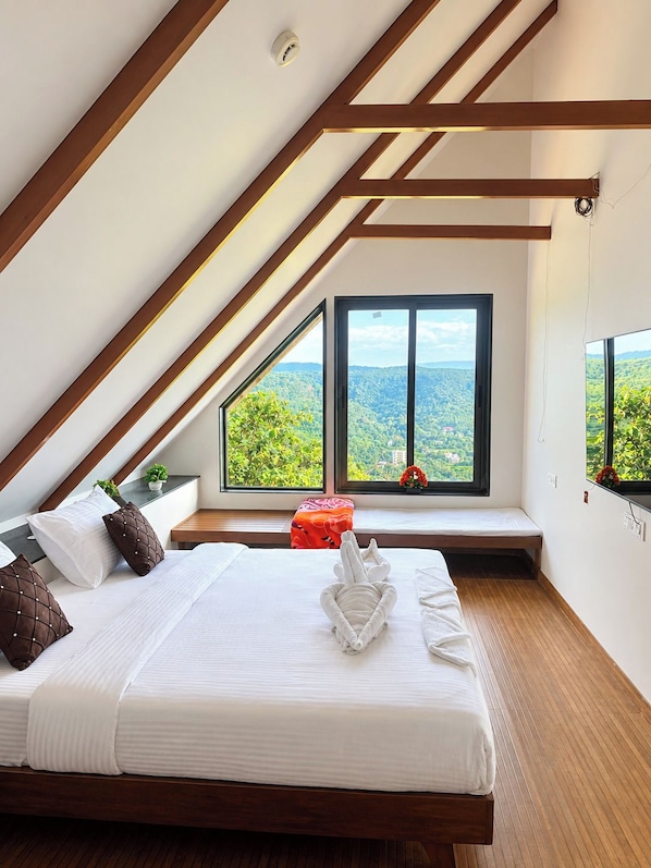 Un dormitorio con techo inclinado, un ventanal con vistas a la vegetación, una cama con almohadas y una mesita de noche.
