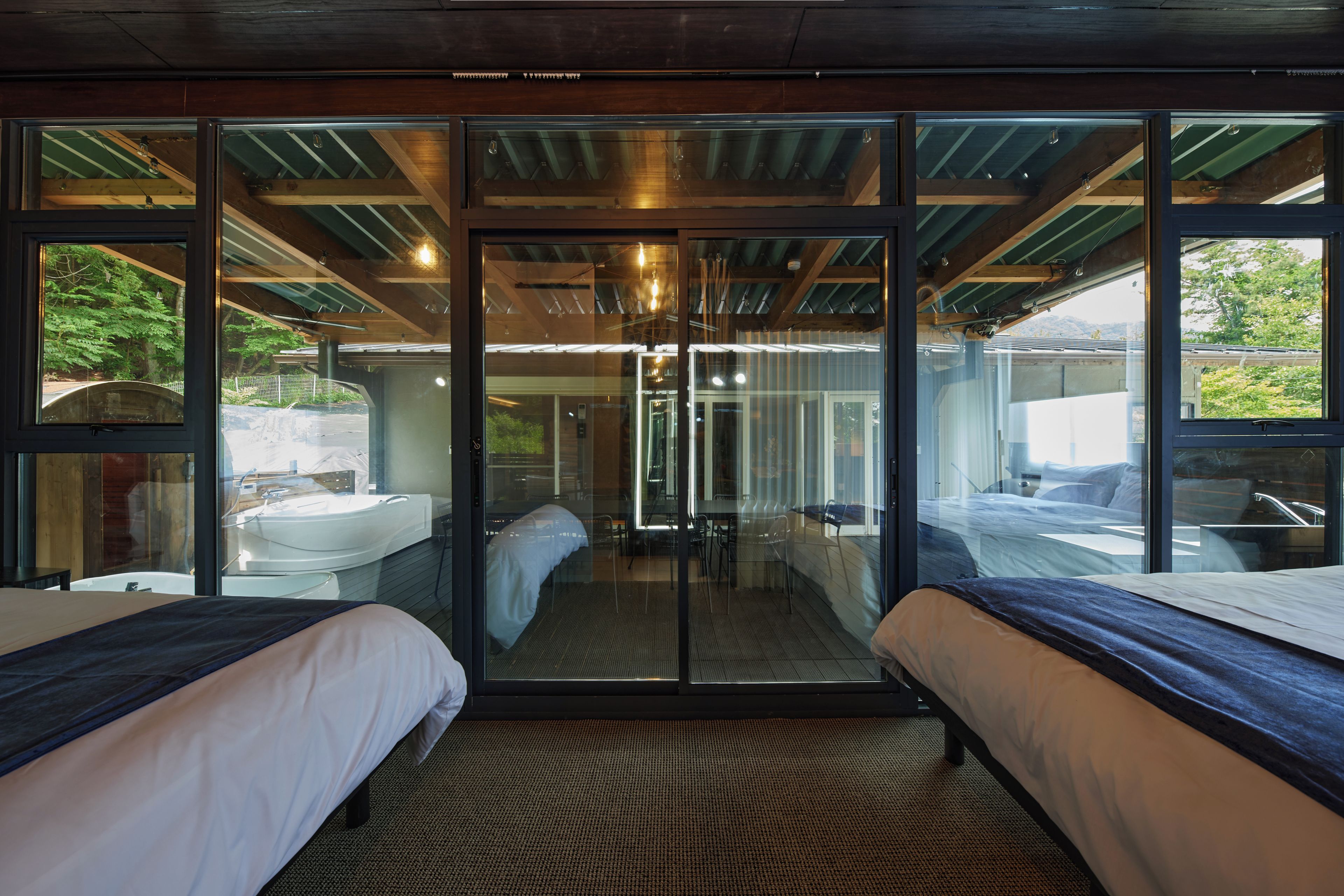 A modern hotel room with two beds, a glass door leading to a balcony, and a view of the outdoors.