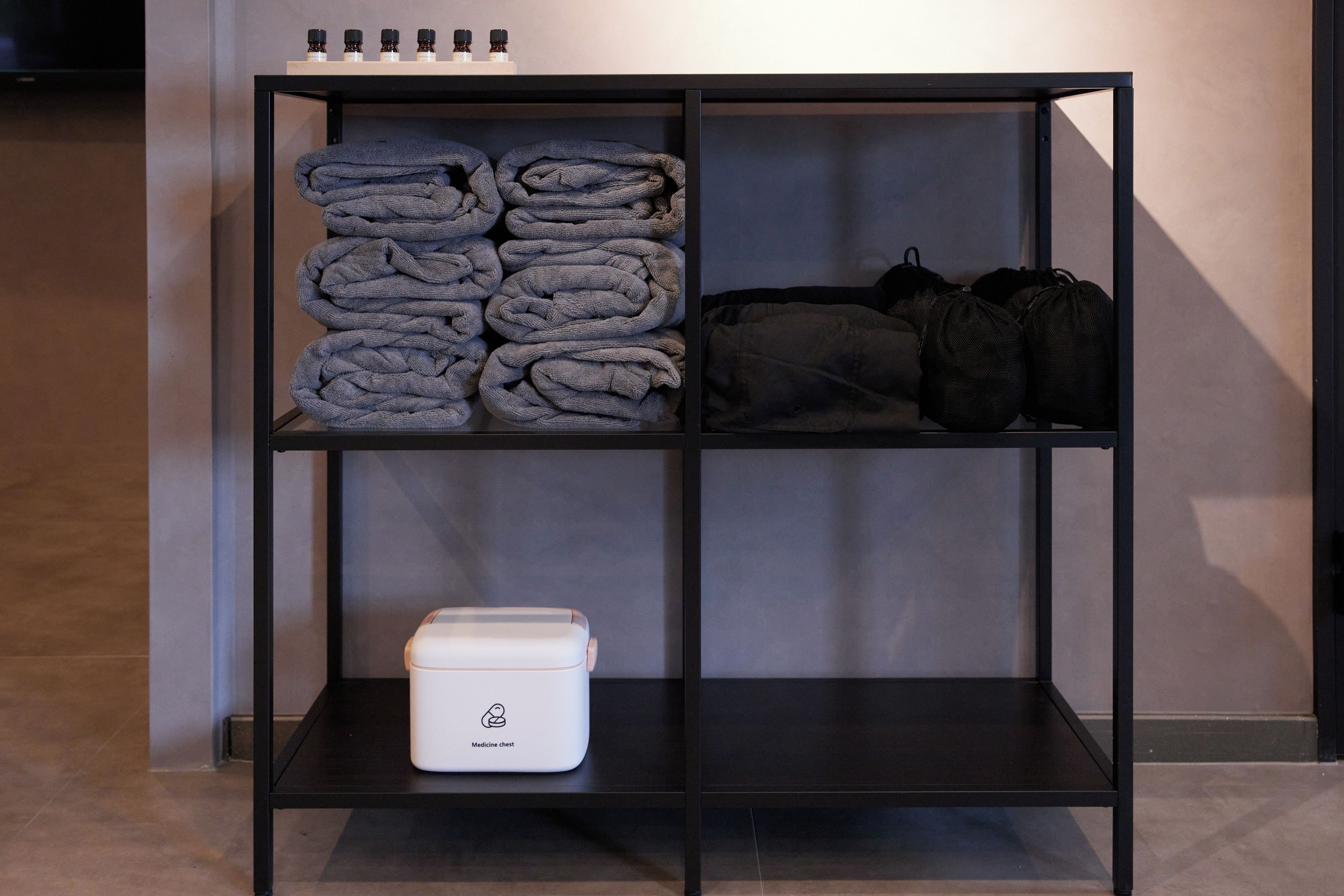A black metal shelf unit with folded towels, a medicine chest, and a pair of black bags.