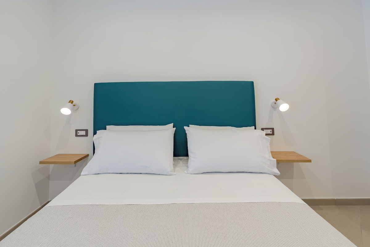 A bed with white bedding, a teal headboard, and two bedside tables with wall-mounted lamps.