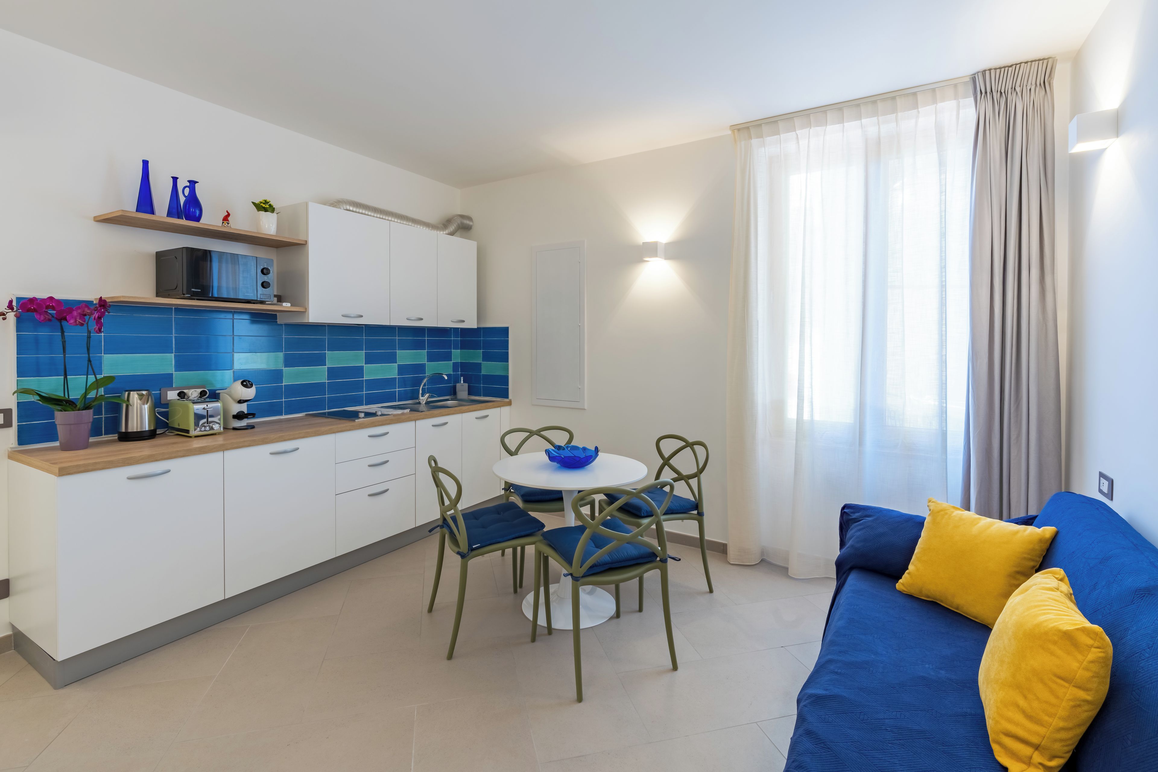 Una sala de estar moderna con un sofá azul y cojines amarillos, un comedor con una mesa redonda y sillas, y una cocina con azulejos azules y armarios blancos.