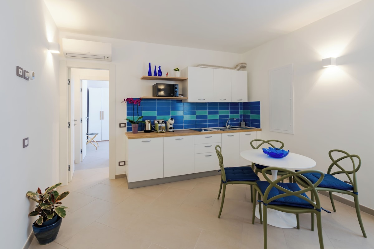 A modern kitchen with white cabinets, a blue tiled backsplash, and a dining area with green chairs and a white table.
