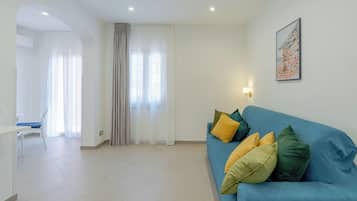 A modern living room with a blue sofa, a dining table, and a chair. There is a painting on the wall and a window with curtains.