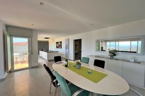 A modern dining area with a white oval table, teal chairs, and a view of the sea through large windows. - Holiday home in Roses with swimming pool (Roses)