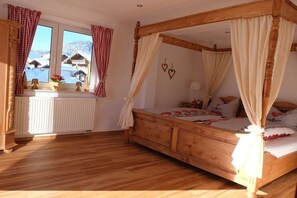 A wooden four-poster bed with a canopy, a white radiator, and a view of snow-covered houses through a window with red and white checkered curtains. - Allgäu Hearts 1 and 2 (Kornau\/Oberstdorf)
