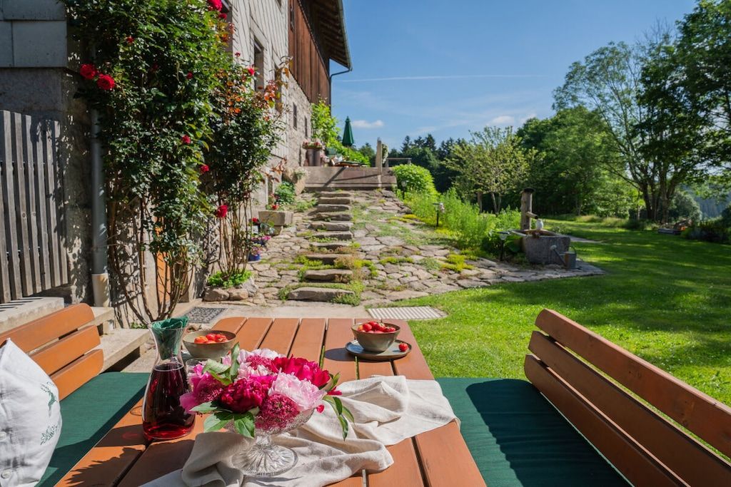 A wooden bench with a table set for dining, a stone pathway leading to a building, and a lush garden with various plants and flowers.