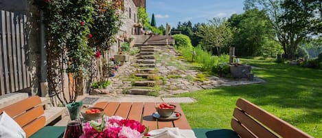 A wooden bench with a table set for dining, a stone pathway leading to a building, and a lush garden with various plants and flowers.