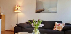 A living room with a black sofa, a vase with flowers, a lamp, and a painting on the wall. - Holiday apartment with 1 bedroom (Langenargen)