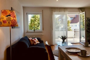 A living room with a sofa, a table with a vase, and a window with a view of a tree and buildings. - Holiday apartment with 1 bedroom (Langenargen)