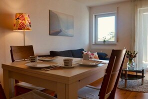 A dining area with a table set for two, a sofa, a lamp, and a window with a view. - Holiday apartment with 1 bedroom (Langenargen)
