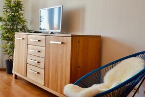 A wooden cabinet with drawers and a television on top, a blue chair with a white cushion, and a potted plant. - Holiday apartment with 1 bedroom (Langenargen)