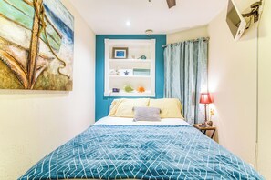 A bedroom with a bed, a nightstand, a wall-mounted shelf with decorative items, and a painting. - Galveston Condo (Galveston)