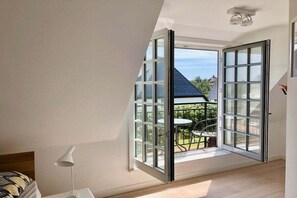 A room with a large window, a bed, a white lamp, and a view of a house and greenery outside. - Holiday home in Hörnum with swimming pool (Hörnum)