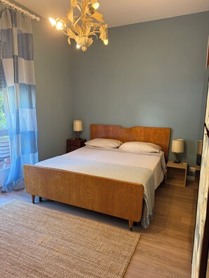 Une chambre à coucher avec un lit en bois, deux tables de chevet avec des lampes, un luminaire suspendu et une vue sur la verdure à l’extérieur.