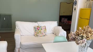 Uma sala de estar com um sofá branco, uma poltrona amarela, uma mesa branca e um vaso com flores secas.