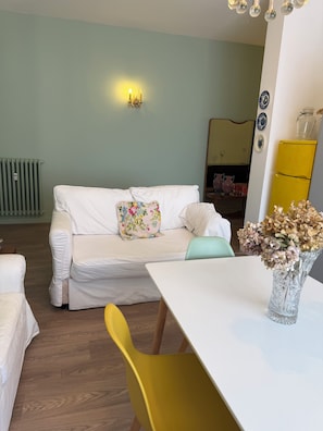 Un salon avec un canapé blanc, un fauteuil jaune, une table blanche et un vase contenant des fleurs séchées.