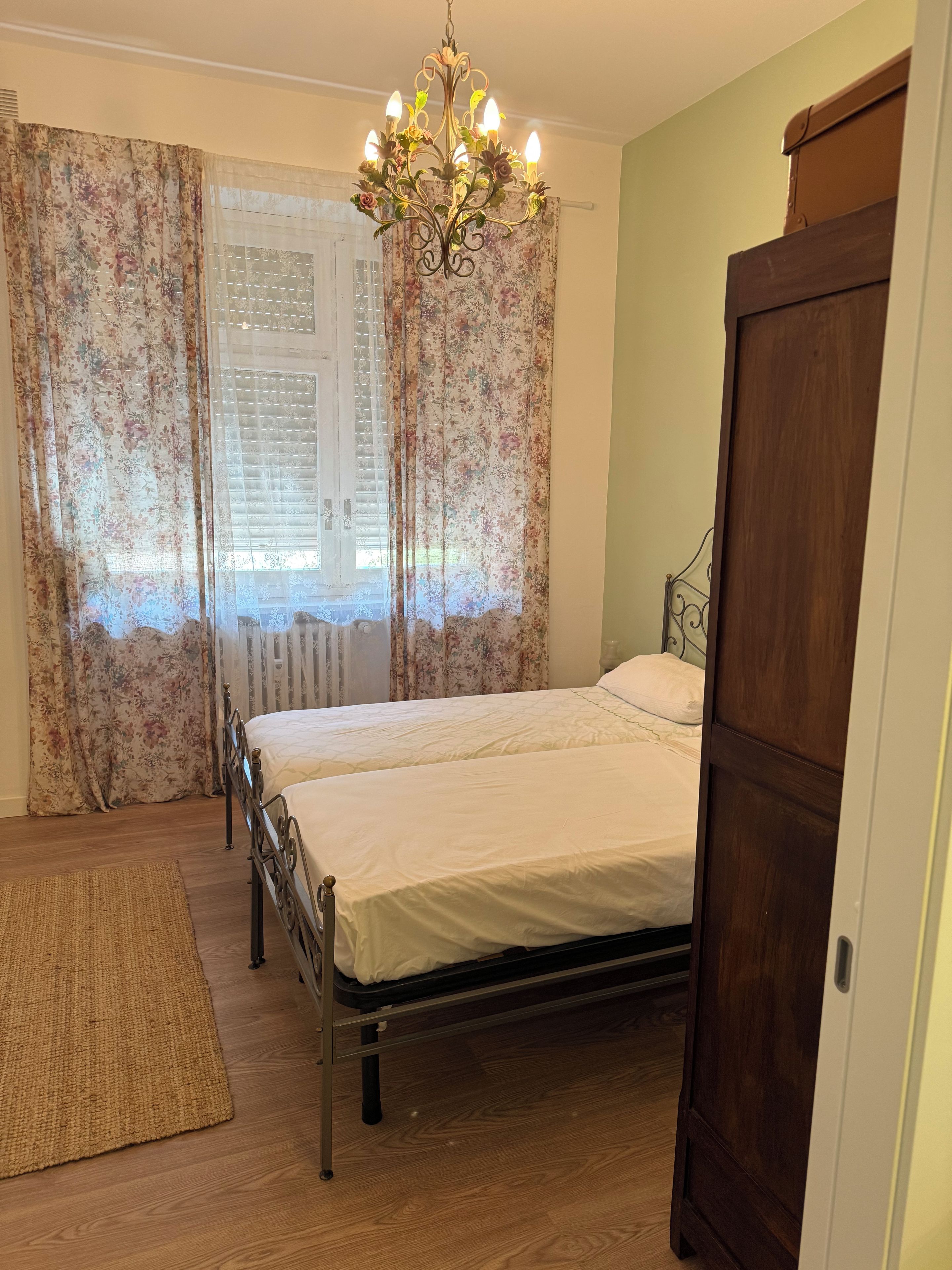A bedroom with a double bed, a wooden wardrobe, a chandelier, and floral curtains.