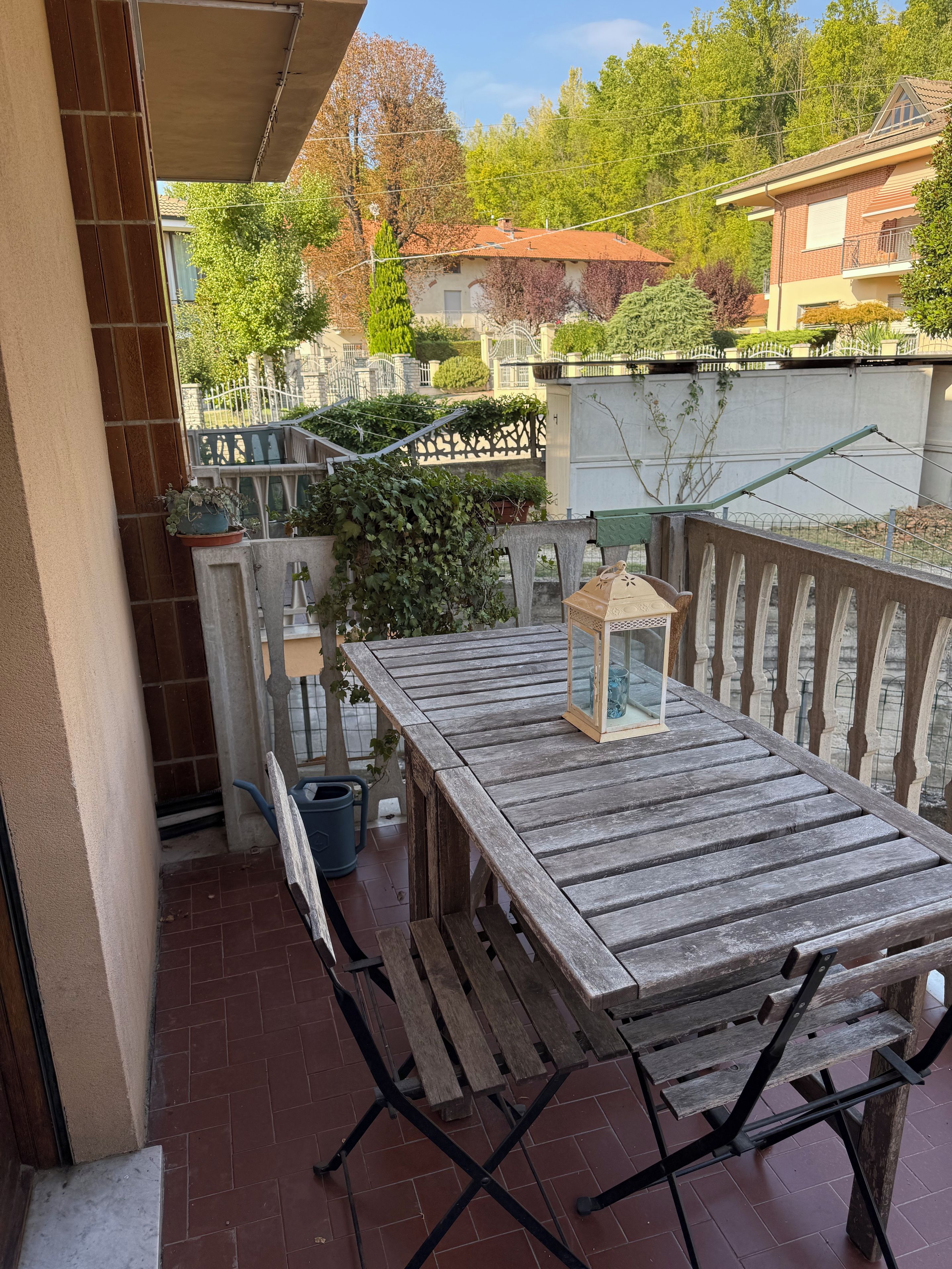 Ein Holztisch mit einer Laterne auf einem Balkon mit Blick auf einen Garten und Nachbarhäuser.