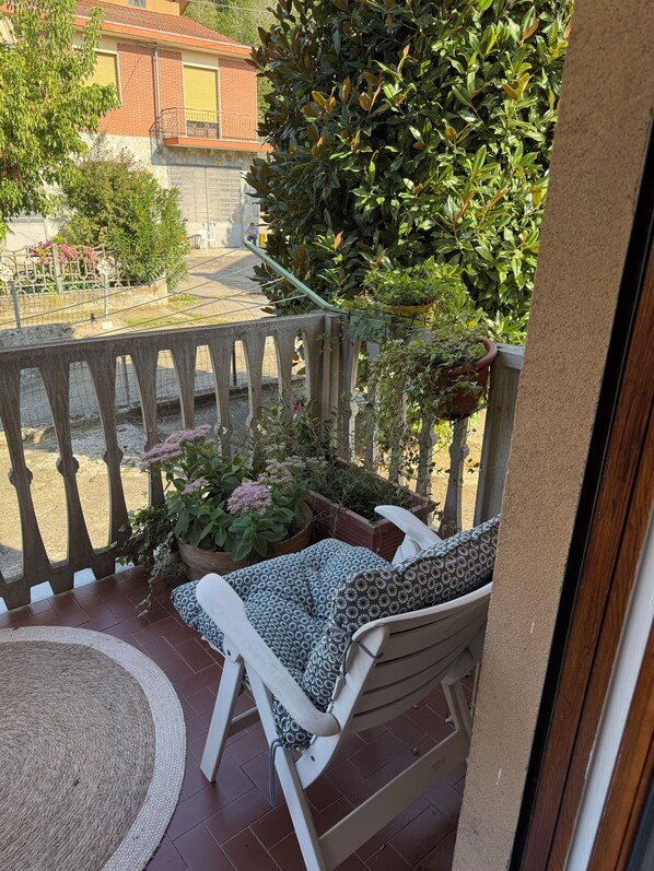 Un balcon avec une chaise blanche et un coussin à motifs, entouré de plantes en pot et offrant une vue sur un immeuble.