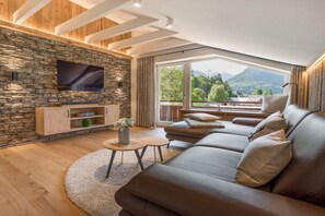 Una sala de estar moderna con una pared de piedra, un televisor de pantalla plana, un sofá y vistas a montañas a través de amplios ventanales.