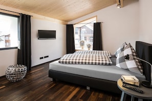 A modern bedroom with a bed, a TV, a window with curtains, and a small table with books and a remote. - Holiday apartment with 2 bedrooms (Oberstdorf)