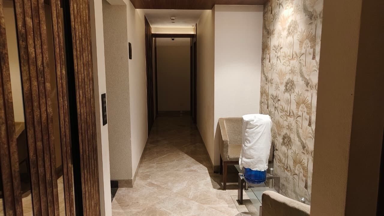 Ein schmaler Flur mit einem Stuhl, einem Tisch, einer Tür mit vertikalen Jalousien und einer Wand mit Muster.
