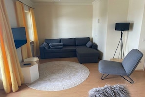 Ein Wohnzimmer mit einer Eckcouch, einem einzelnen Sessel, einem runden Teppich, einem kleinen Beistelltisch, einer Stehlampe und einem Fernseher.