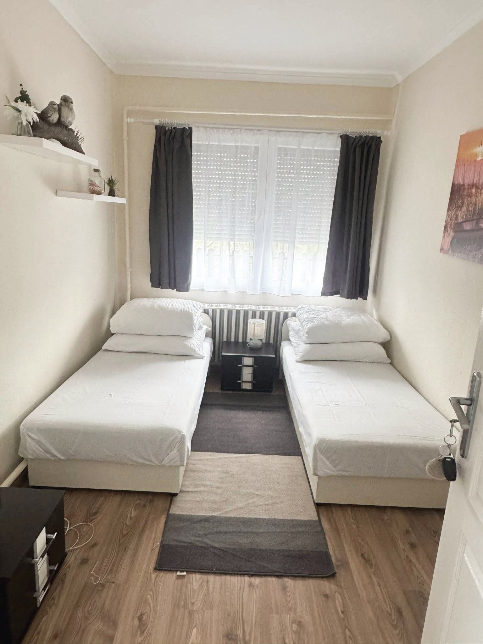 Une petite pièce, de taille modeste, comprenant deux lits simples, un radiateur, une fenêtre avec des rideaux, une petite étagère avec des objets décoratifs et une porte avec un porte-clés.
