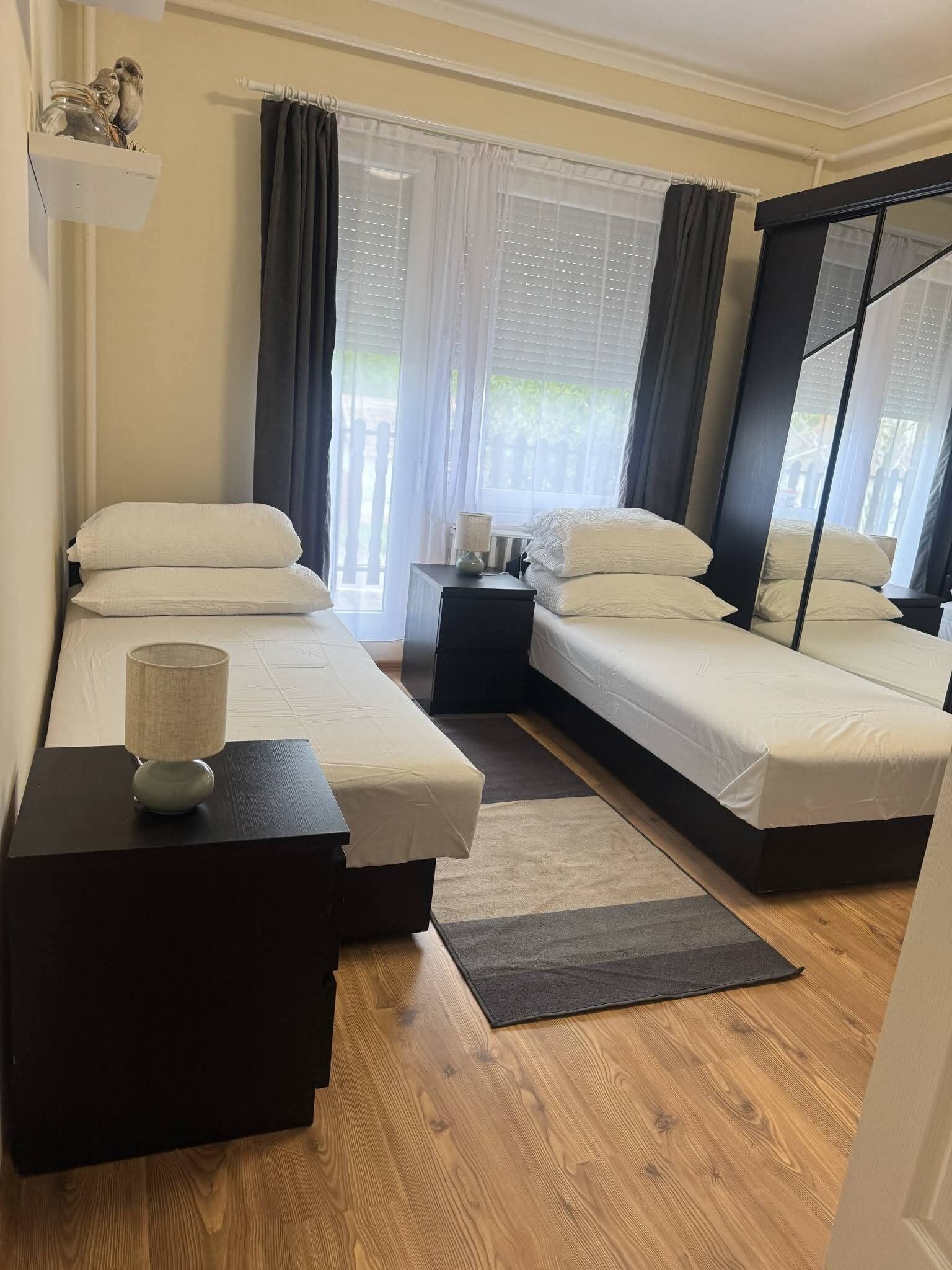 Ein Schlafzimmer mit zwei Betten, einem Nachttisch mit Lampe, einem kleinen Tisch mit Vase und einem Fenster mit Vorhängen.