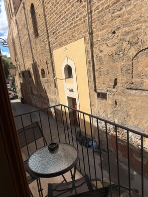 Ein Balkon mit Metallgeländer, ein runder Tisch mit einem Stuhl und Blick auf ein historisches Gebäude mit Rundbogenfenstern und einer Tür.
