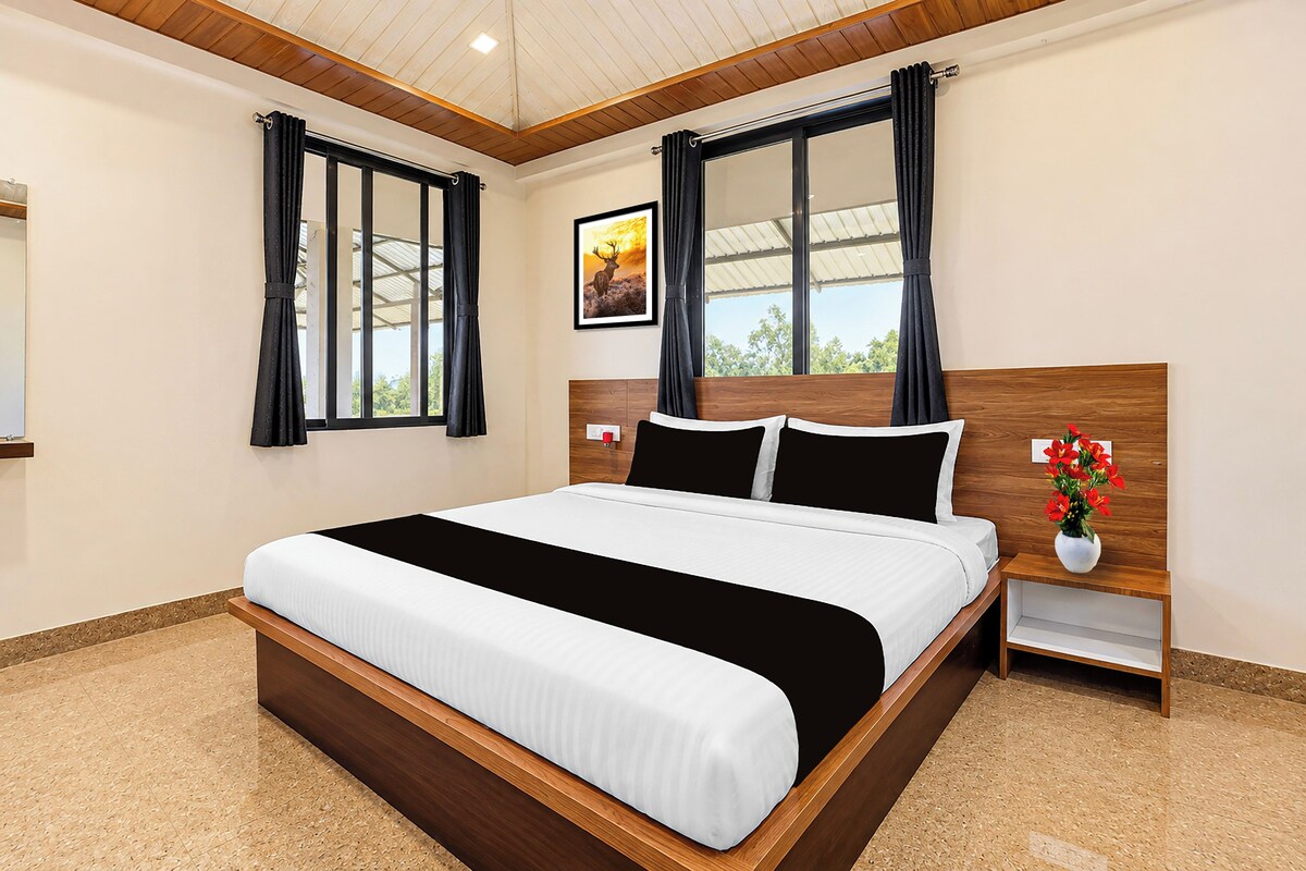 Una habitación de hotel moderna con una cama grande, cabecera de madera, ropa de cama blanco y negro, una mesita de noche con un florero y una ventana con cortinas.